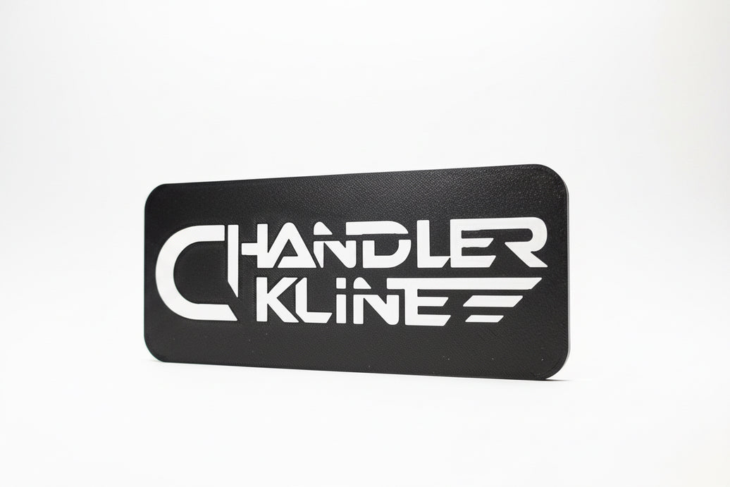 A black rectangular sign or patch with rounded corners featuring the stylized white text 'CHANDLER KLINE' against a dark background.