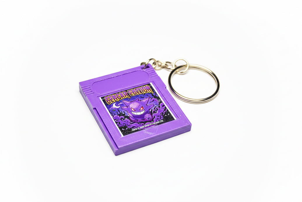 A purple Gengar-themed keychain designed to look like a classic Game Boy game cartridge, featuring a gold split ring on a textured black background.