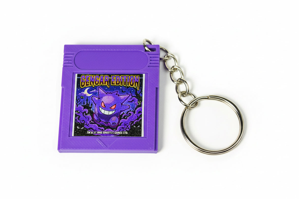 A purple Gengar Edition game cartridge keychain with a metal split ring, displayed on a dark textured fabric background.